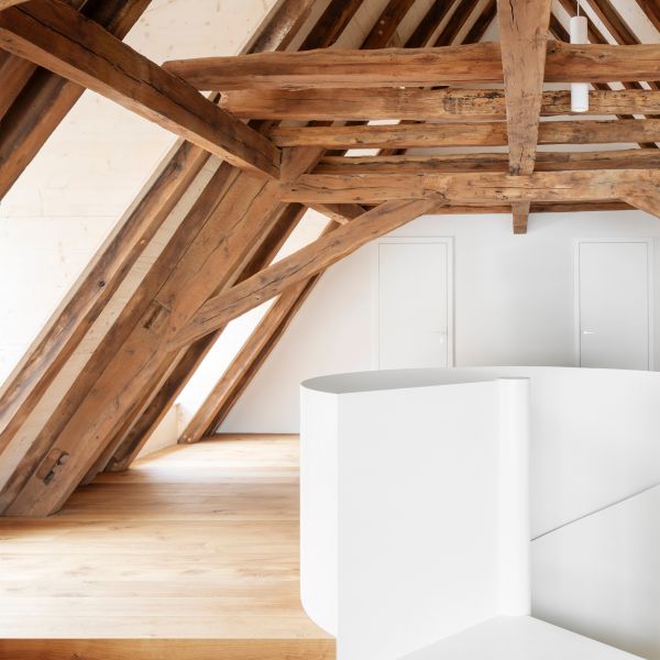 Dachboden mit sichtbaren, massiven Holzbalken und weißer, geschwungener Wand im Vordergrund.