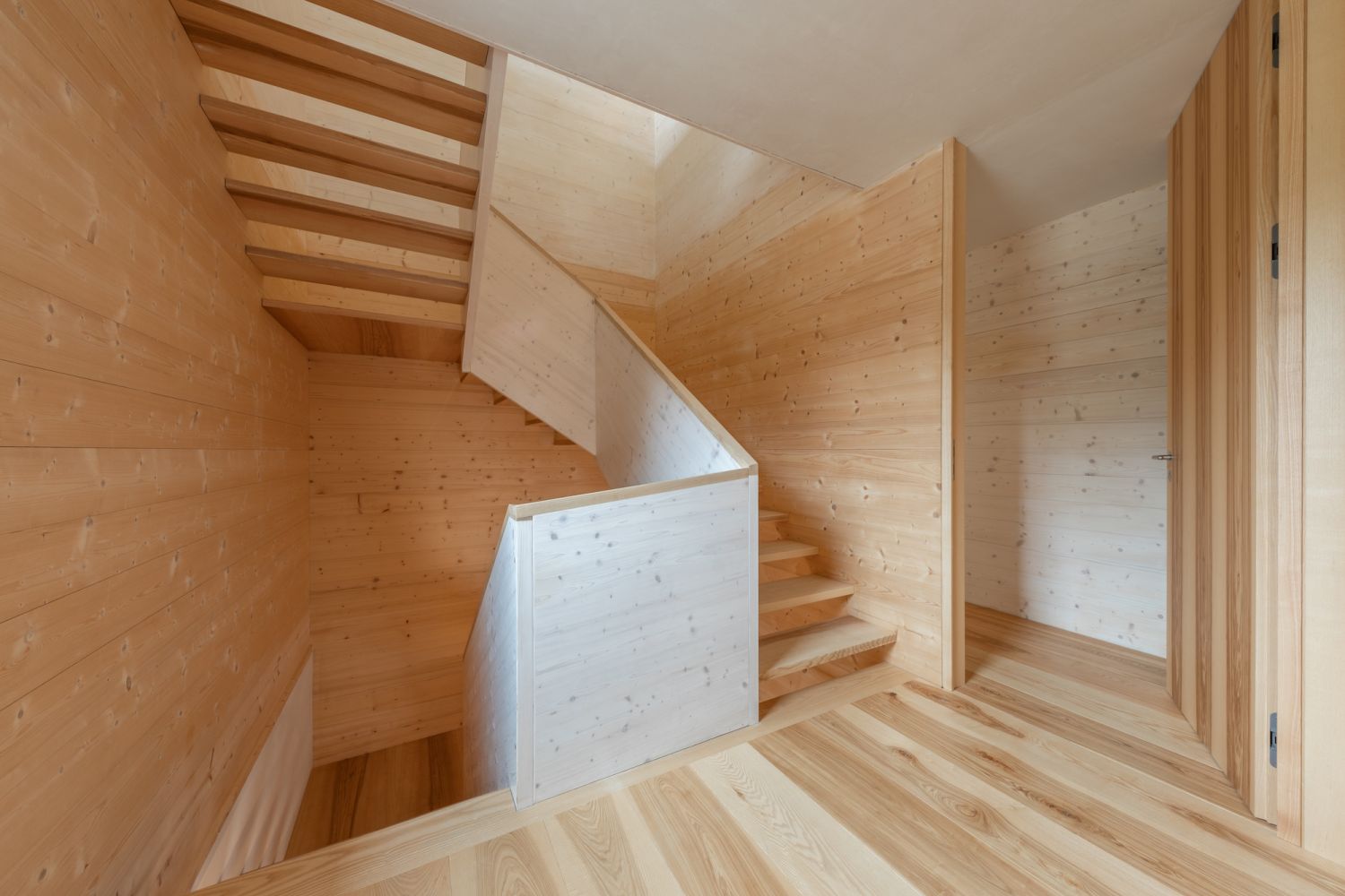 Stairwell made of ash wood with straight stairs and wooden railing, walls and ash wood floor also made of wood.