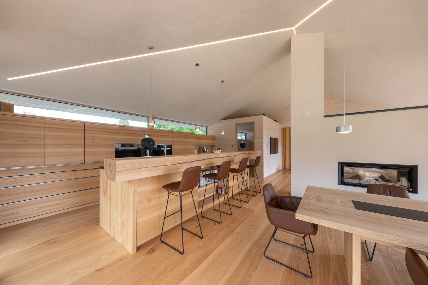 Modern open living area with ash wood flooring, long wooden counter with four brown bar stools, and dining table with brown chairs.