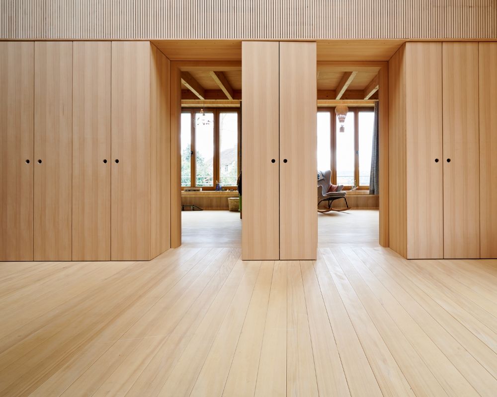 Piece avec sol en bois clair et trois grandes portes coulissantes en bois, derriere lesquelles une fenetre avec vue sur l’exterieur et deux fauteuils.