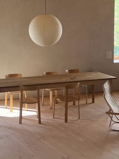 Une salle à manger dans une ferme rénovée avec de larges lames en sapin Douglas. Elle est équipée d’une table en bois simple, de chaises en bois modernes et d’une grande lampe ronde blanche.
