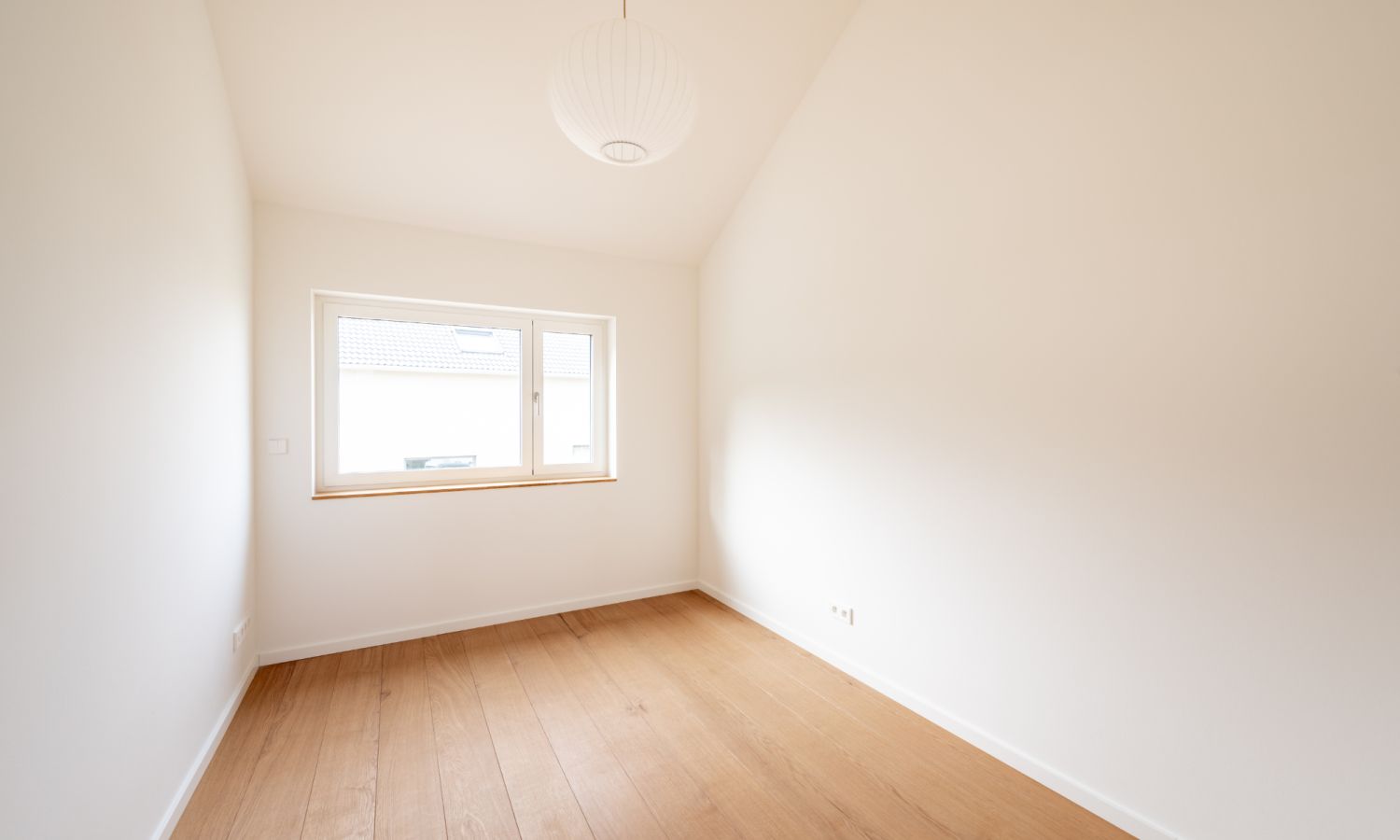 Une pièce vide avec des murs blancs, un parquet en chêne sur toute la longueur et une suspension blanche et moderne. Au fond de la pièce, une fenêtre laisse entrer la lumière naturelle.
