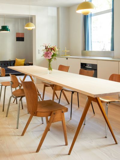 Salle à manger avec une longue table blanche; chaises variées; bouquet de fleurs dans un vase; grandes fenêtres avec rideaux bleus