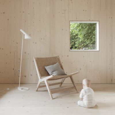 Enfant en grenouillere blanche assis sur un sol en bois clair devant une chaise en bois avec coussin gris, a cote d’une lampe sur pied blanche et d’une fenetre avec vue sur la verdure.
